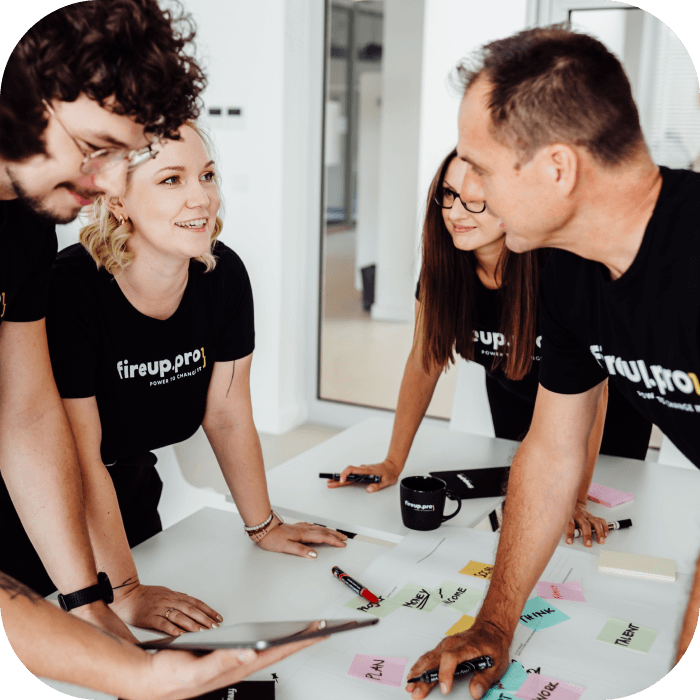 The team, dressed in black t-shirts with the fireup.pro logo, is discussing managed IT services in a meeting. A developer with glasses is presenting his ideas on a tablet, while colorful notes with key client priorities are spread across the table.