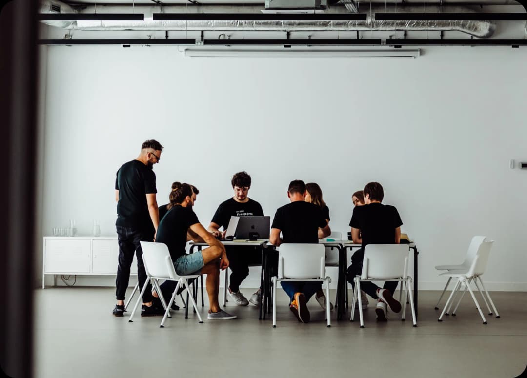 The IT services refactoring process is being discussed in a company workspace by a large team of developers. We're combining their professional experience and knowledge to collectively brainstorm the benefits for the client, the tech stack, and business requirements. Sitting on white chairs, we're crafting the strategy together.