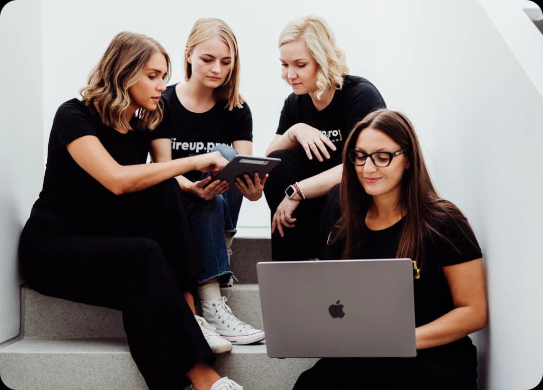 Four women from the fireup.pro team, sitting on the stairs with laptops and tablets, are planning interface designs for our clients. Rely on their experience gained from international projects – fireup.pro, Your UI/UX design company.