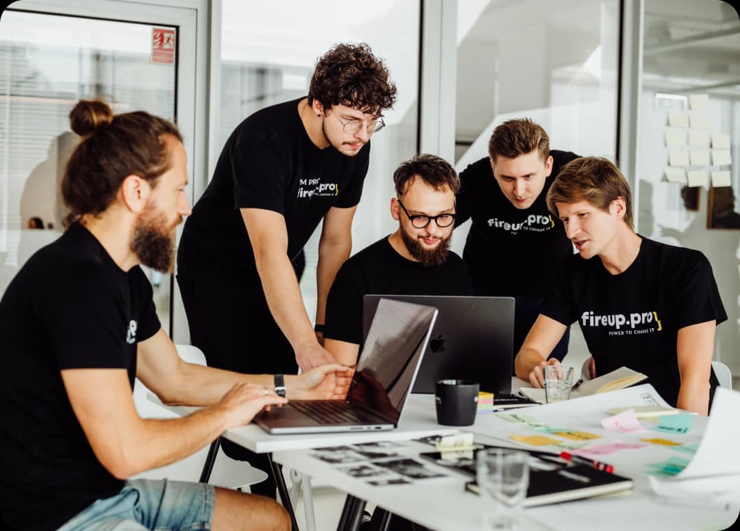 Five developers, using their skills and experience, are sitting around a table planning the backend development strategy and doing notes on mac.