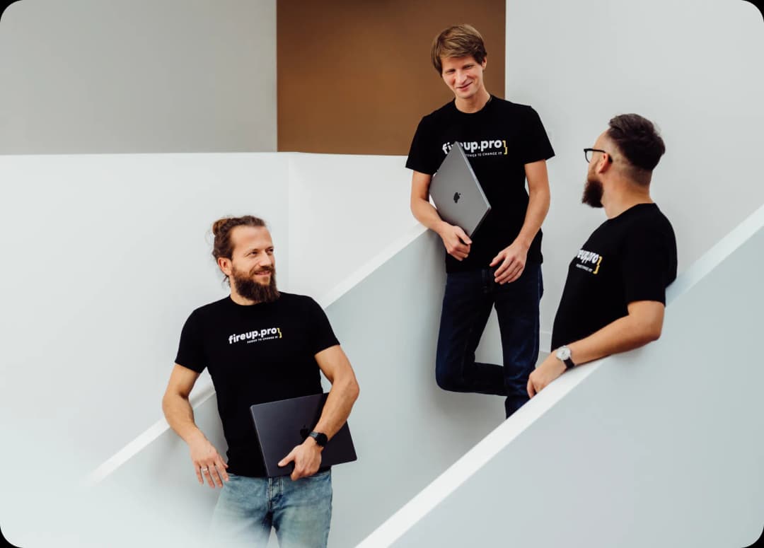 Three developers are standing on minimalist stairs, one holding an Apple laptop, discussing web application development while utilizing all their IT skills. They know why fireup.pro is Your Web Applications Development Company.